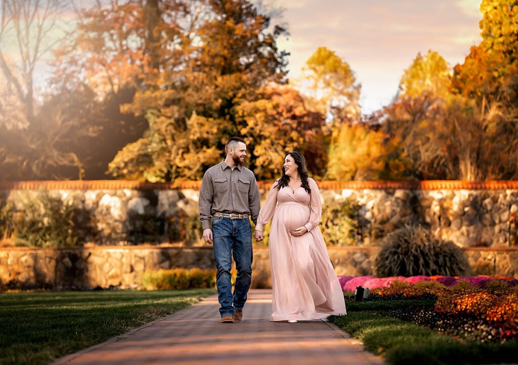 An expecting couple holding hands and walking down a path in the gardens at the biltmore estate.