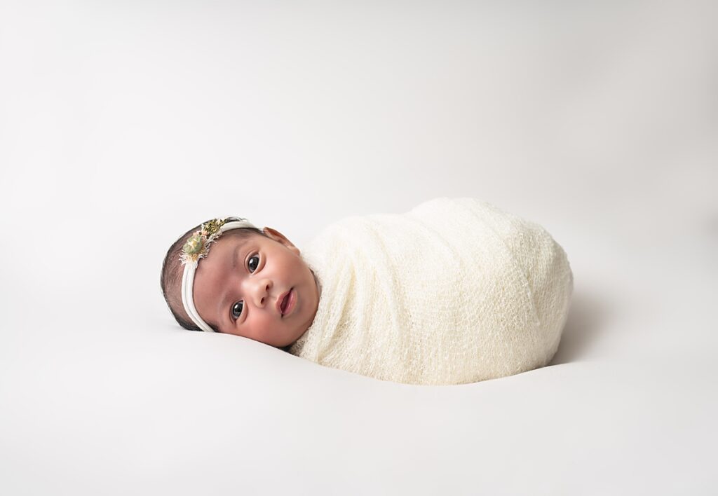 A newborn baby girl wrapped in a white blanket with a flower headband laying on a white blanket very alert. 