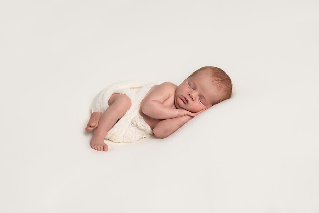 Newborn baby sleeping with hands under his face on a cream blanekt. 