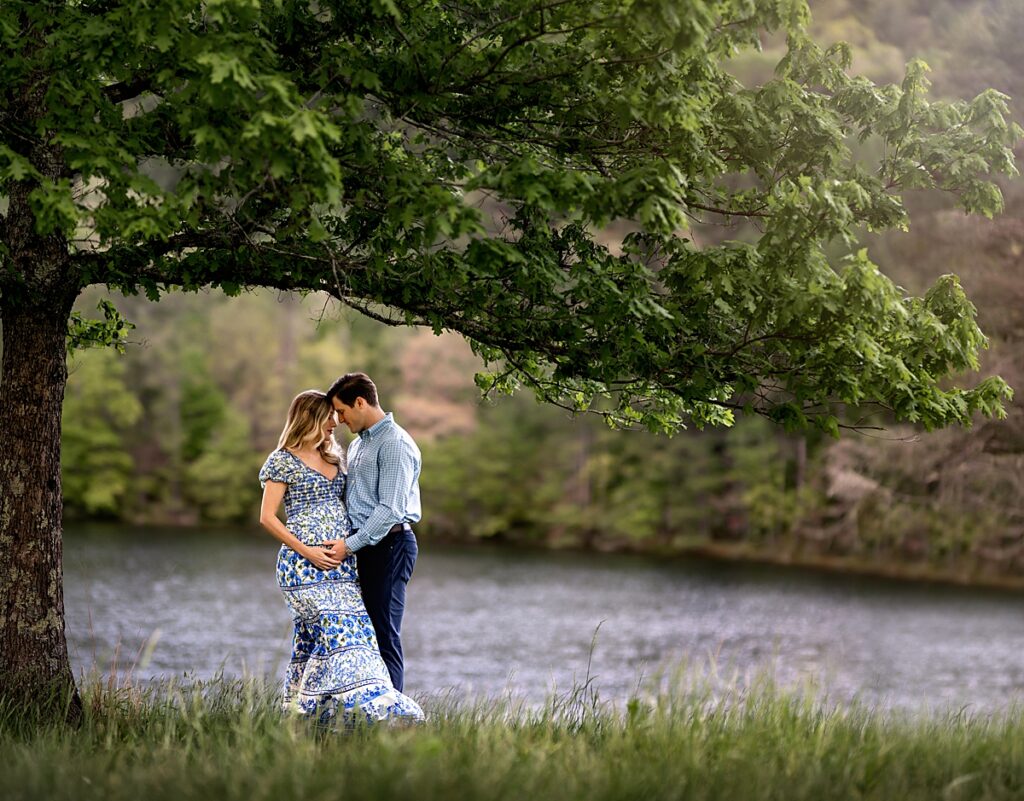 An expecting couple standing under a tree and cuddling by a beautiful lake outside of Asheville NC
