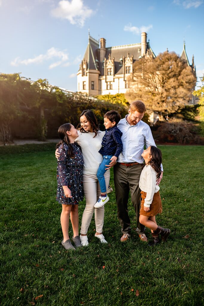 A family of five laughing and cuddling on the lawn beside the Biltmore House in Asheville, NC