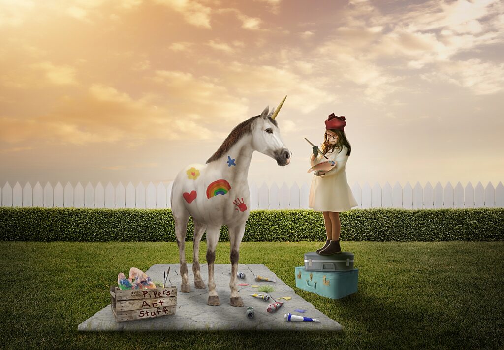 A fantastical image of a little girl standing on suitcases and painting her pet unicorn created by an Asheville Childrens Photographer.