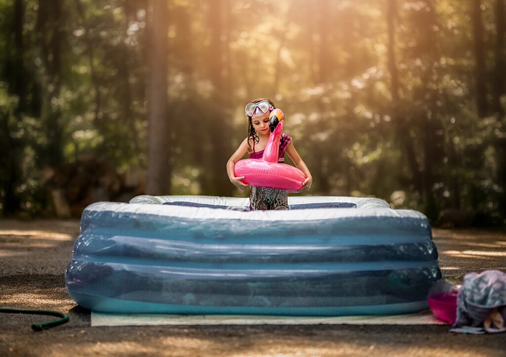A little girl stands in a blow up pool holding a pink flamingo float around her waist.