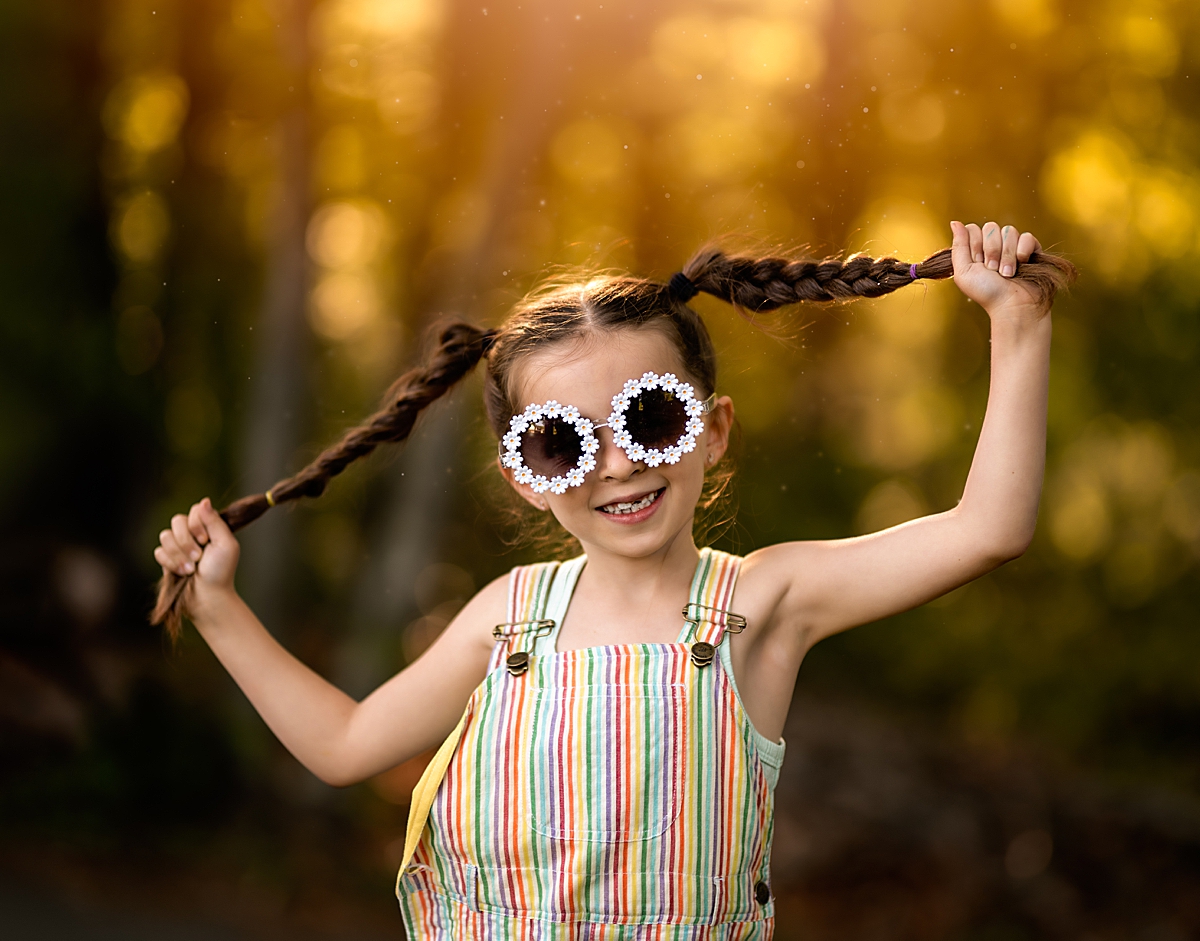 An adorable girl in overalls and sunglasses with flowers around them pulling on her pigtails