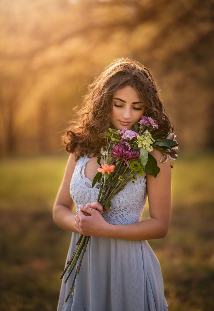 A gorgeous girl in a blue dress holding flowers and making a wish as the sun sets behind her