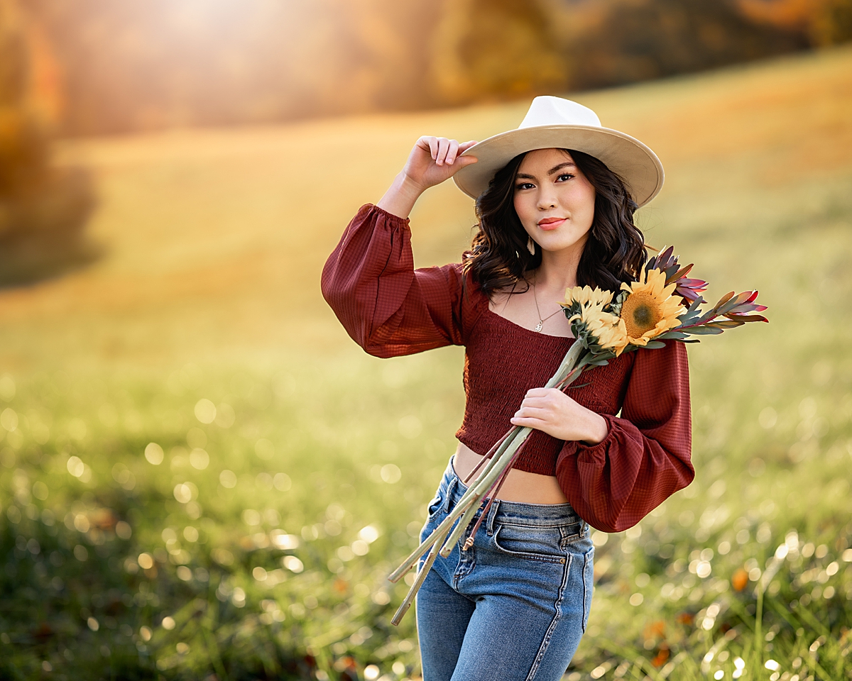 A young girl in a red top and white hat holding sun flowers while standing in sunset during her Asheville Senior Photography session
