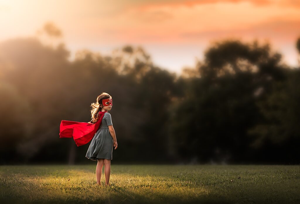 A young girl standing in the sunlight wears a superwoman cape and mask purchased from an Asheville toy Store.