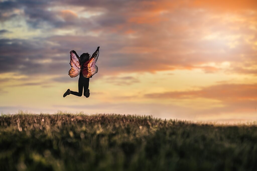 A young girl wearing butterfly wings jumps up in the air as the sunsets behind her. 