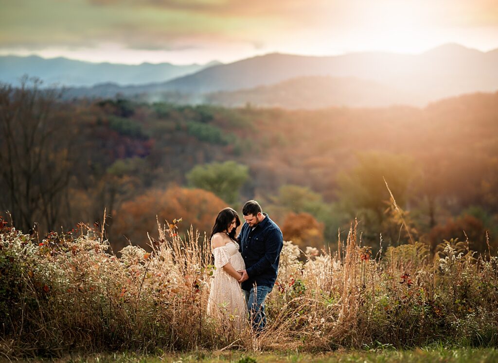 An adorable expecting couple cradling their baby bump while standing in the tall grass at sunset. 
