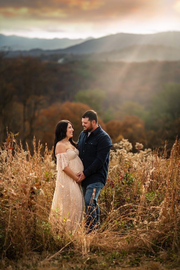 An expecting couple standing in the tall golden grass at sunset for a portrait session with a maternity photographer in Asheville