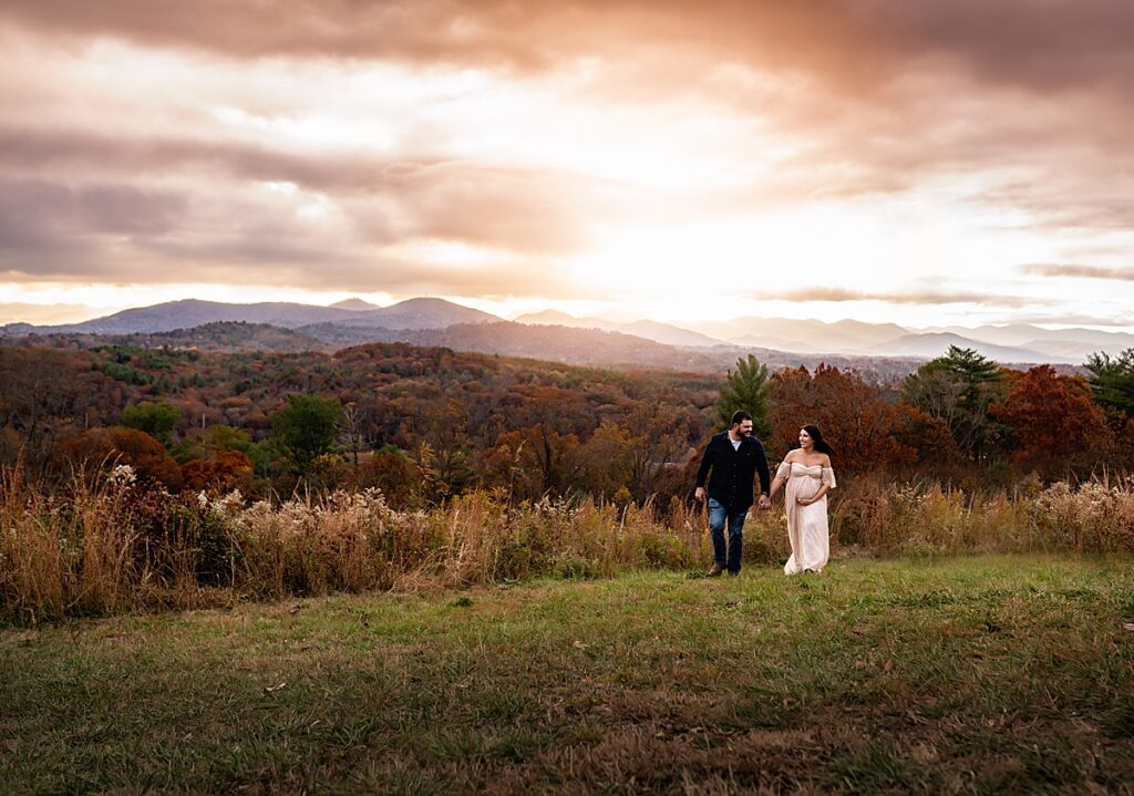 An expecting couple walking across a grass meadow with mountains in the distance near Asheville, NC. 