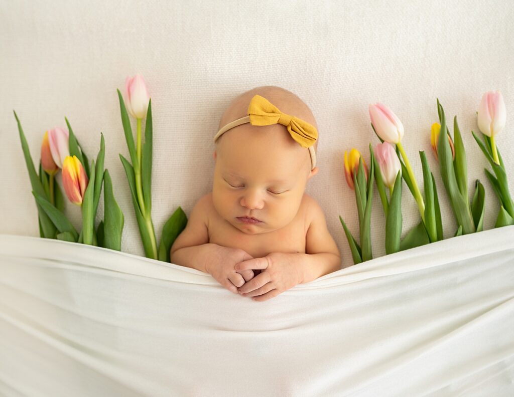 A newborn baby girl with a yellow headband sleeps while surrounded by tulips during her portrait session with An Asheville Studio Newborn Photogapher
