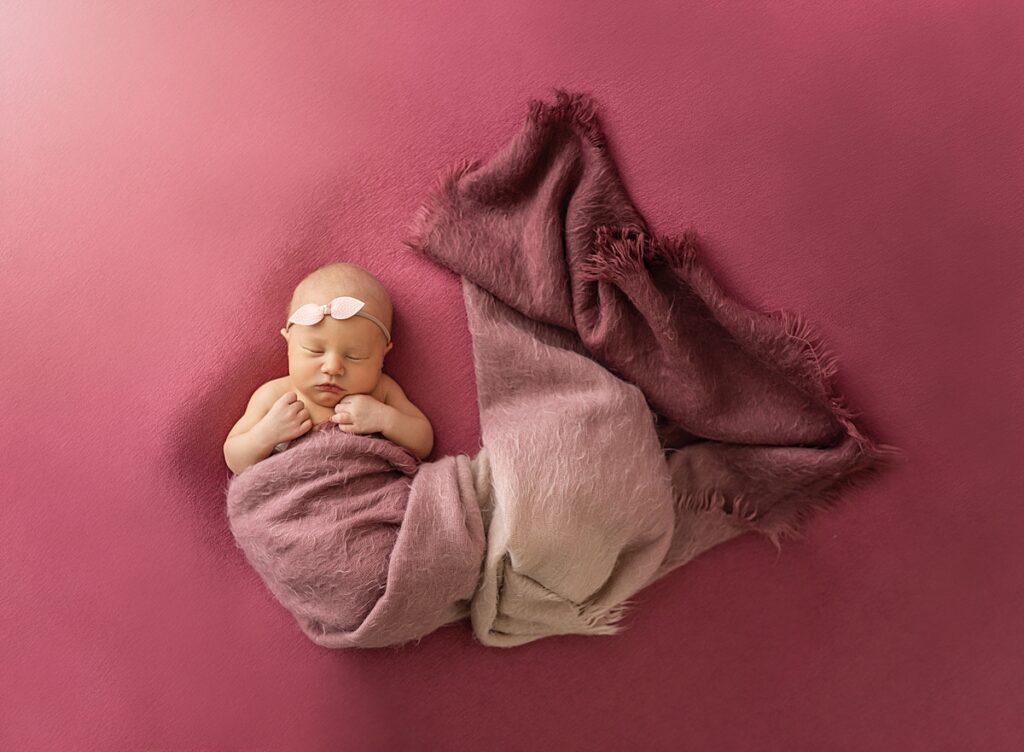 A newborn baby girl wrapped in a dark pink and purple blanket with a pink headband on. 