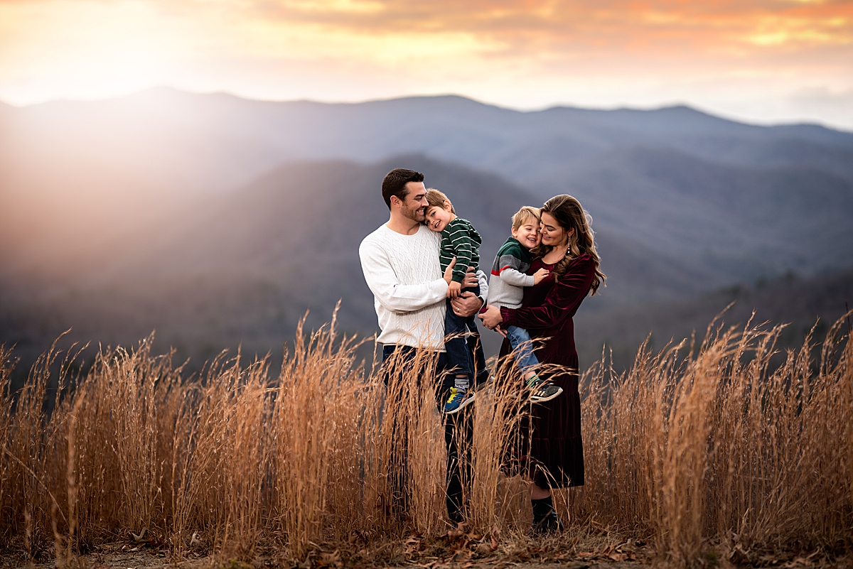 A family of four snuggling in the tall grass with mountains in the distance near Asheville NC