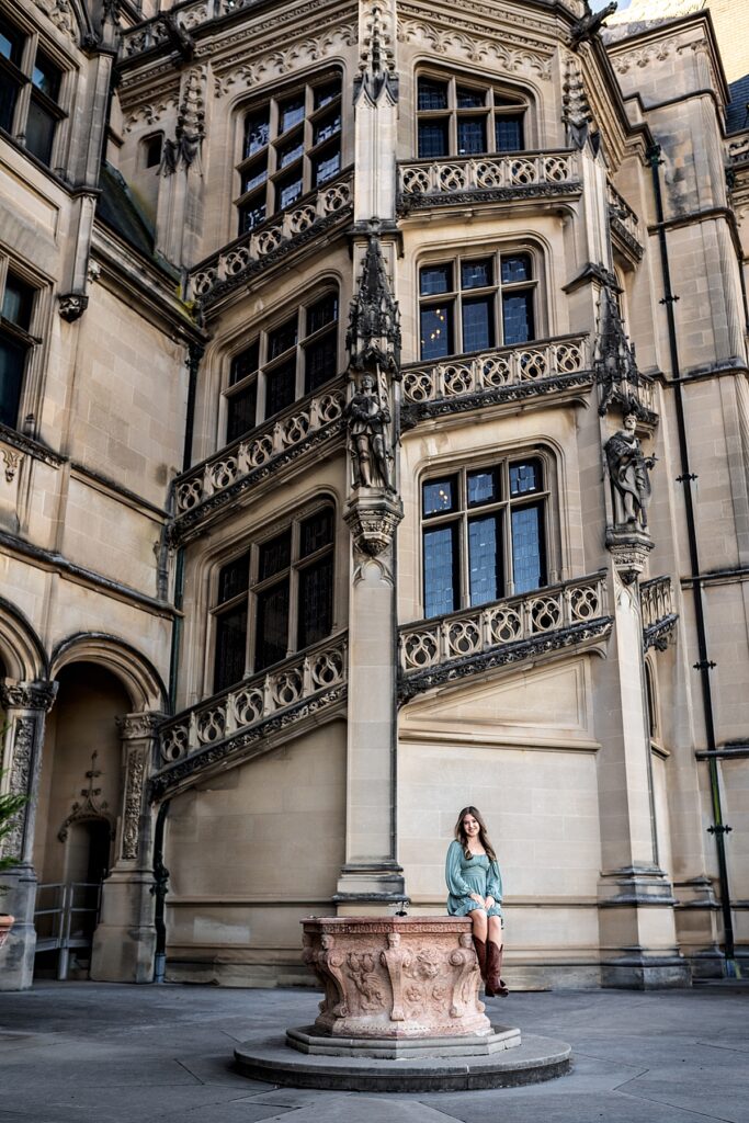 A beautiful girl in a teal dress sits on a fountain outside the Biltmore House during a session with a high school senior photographer in Asheville 