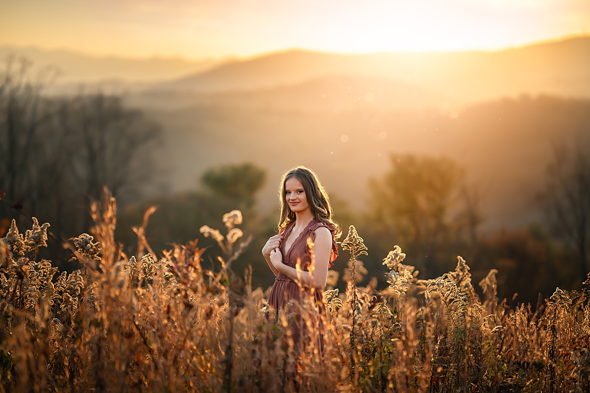 beautiful girl standing in tall grass with the mountains in the distance at her senior photo session in Asheville