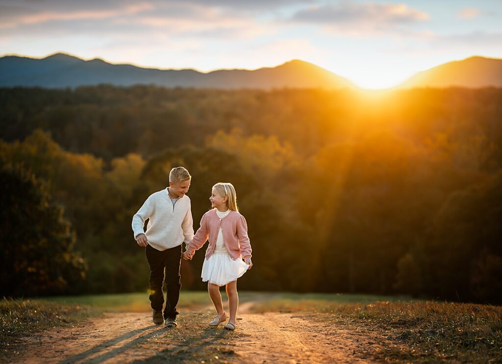 A brother and sister holding hands and running together as the sun sets over the mountains behind them