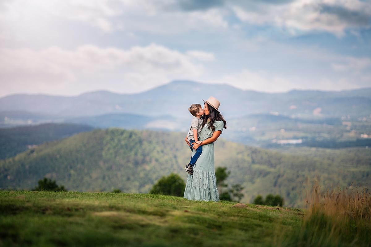 A beautiful mom in a blue dress holding her little boy and kissing his cheek with mountains behind during her session with Asheivilles Best Family Photographer