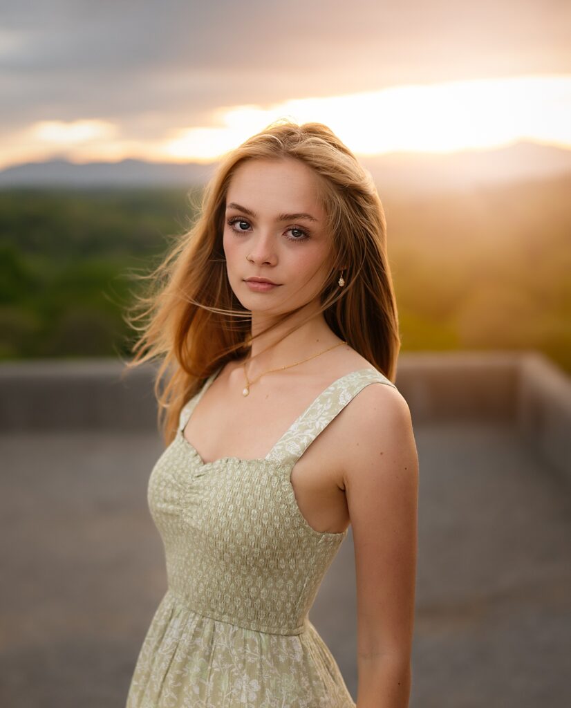 A beautiful young girl in dress stands at the Biltmore Estate with the mountains and sunset behind her. Her hair is blowing in the wind at her senior photos at the Biltmore Estate