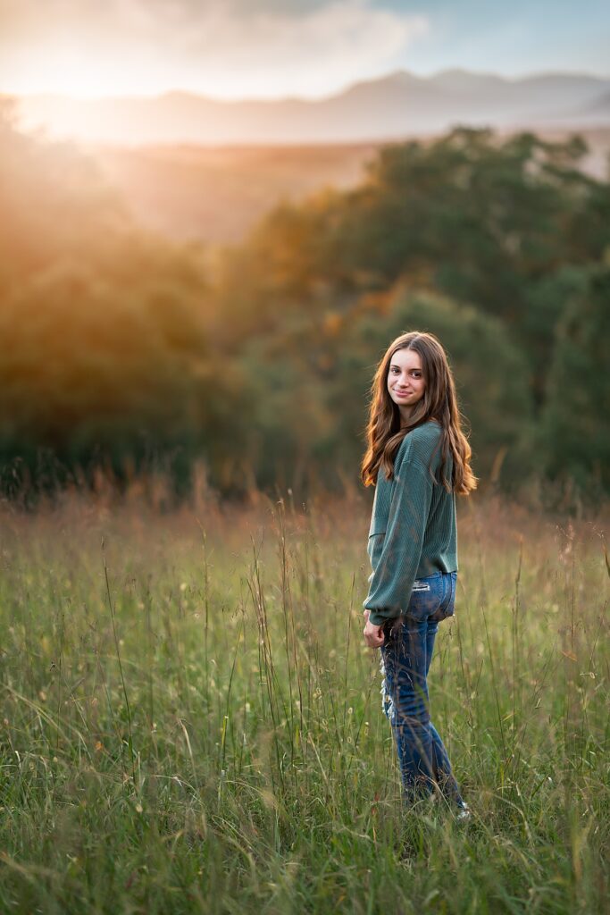 A beautiful, young girl stands in tall grass at sunset with mountains behind her 