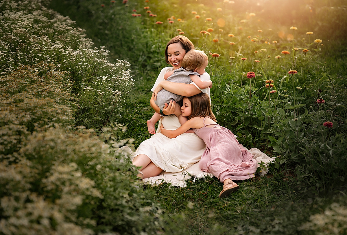 A mom and her two kids snuggle on a blanket in the middle of a flower garden. They are smiling and happy. They are getting artful family photos in Asheville NC