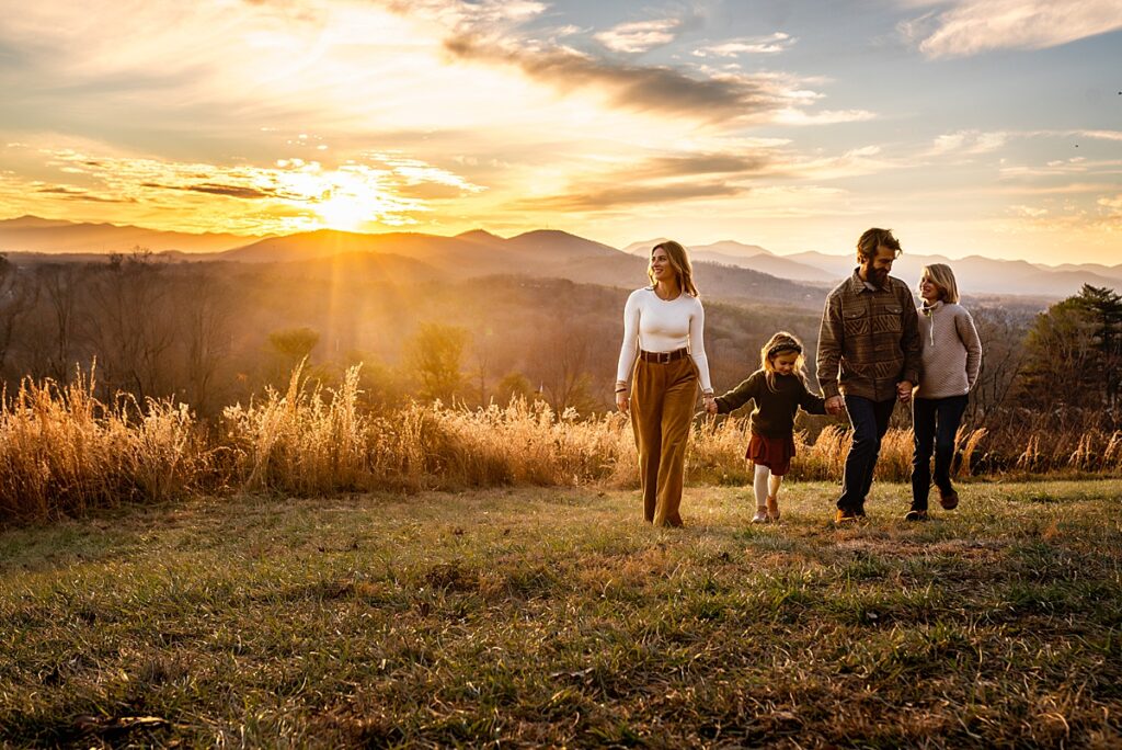 A mom, dad, son and daughter hold hands and walk along a mountain top as the sun sets behind them while getting artistic photos in Asheville, NC