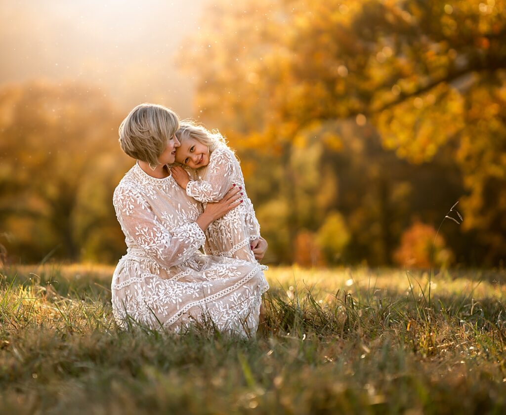 A mom and daughter wearing beautiful white dresses snuggle and smile as the golden sun sets behind them. 