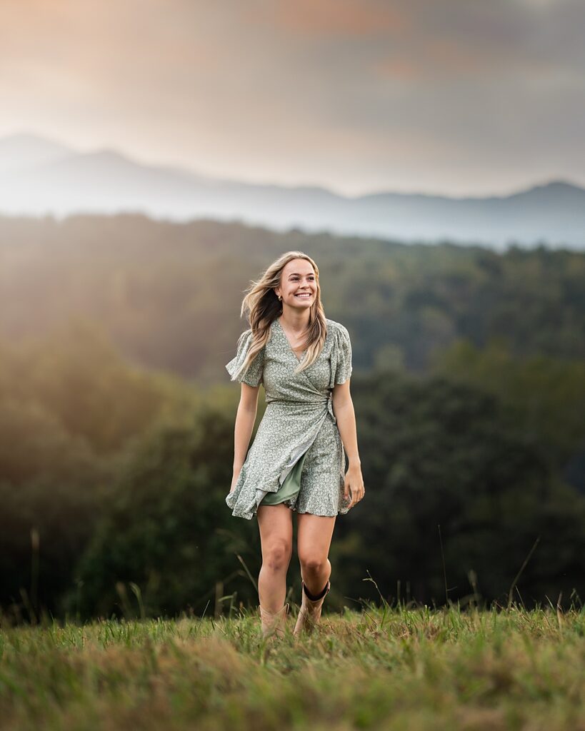 A girl in a green dress and boots walks in the grass with mountains behind her and the wind in her hair 