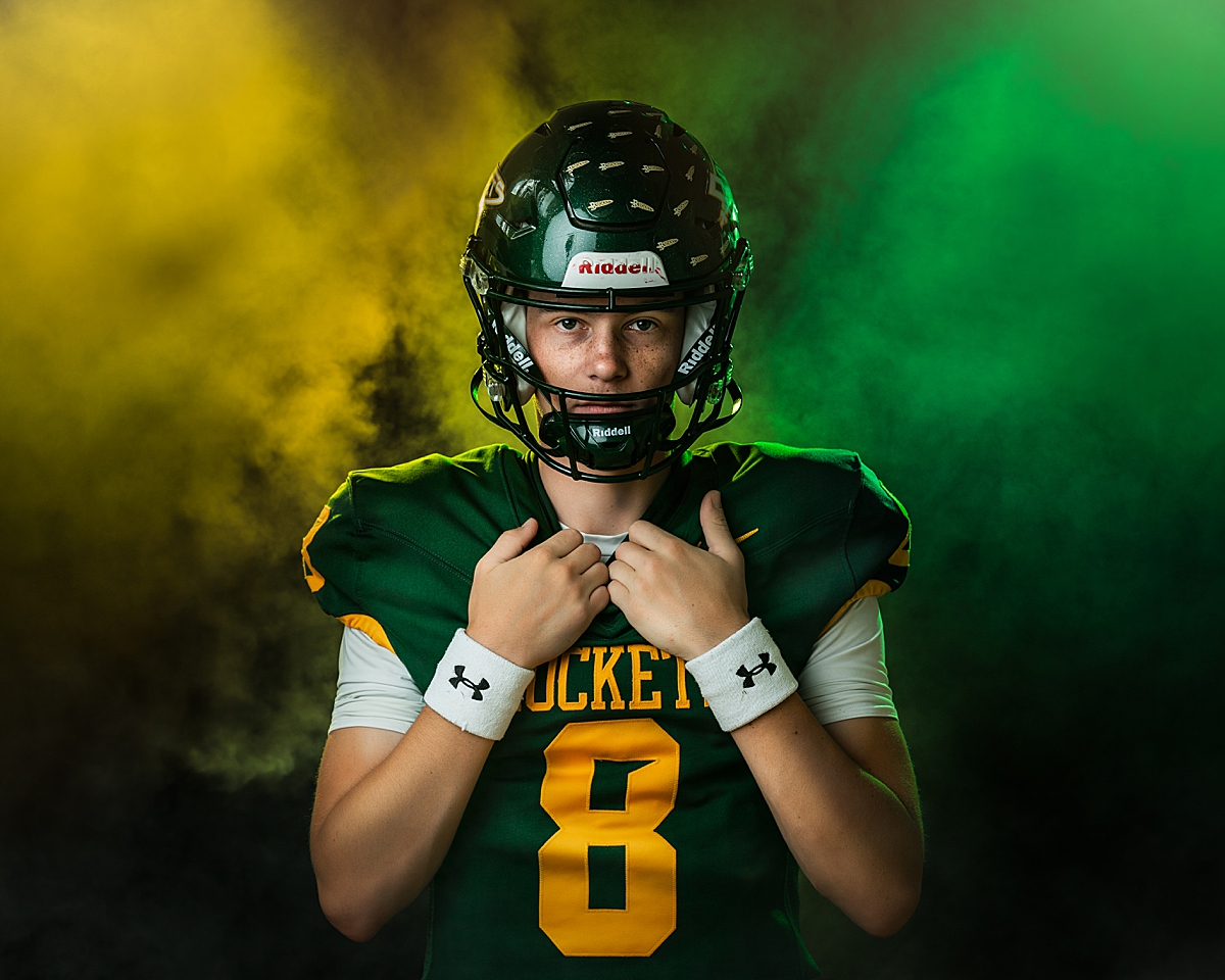 A high school boy in his football uniform standing in green and gold smoke.