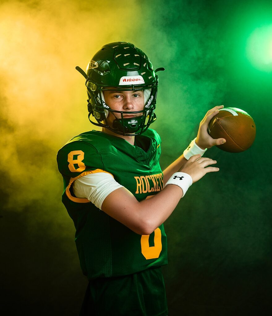 A senior quarterback dressed in his uniform prepared to throw a ball as he stands in green and gold smoke. 