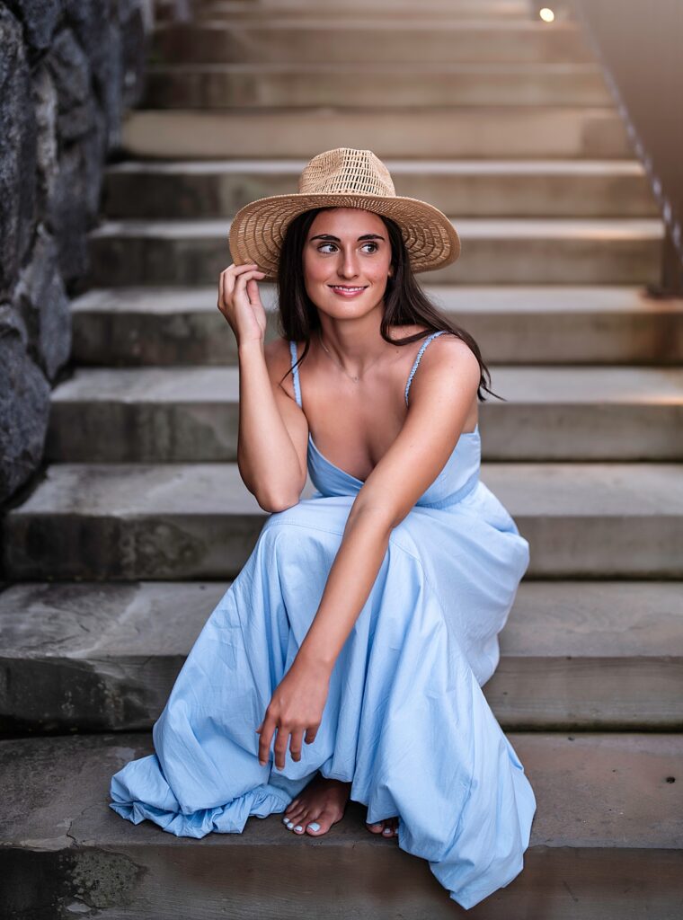 A stunning girl with dark hair wears at hat and blue dress and sits on the steps at the garden at the Biltmore Estate 