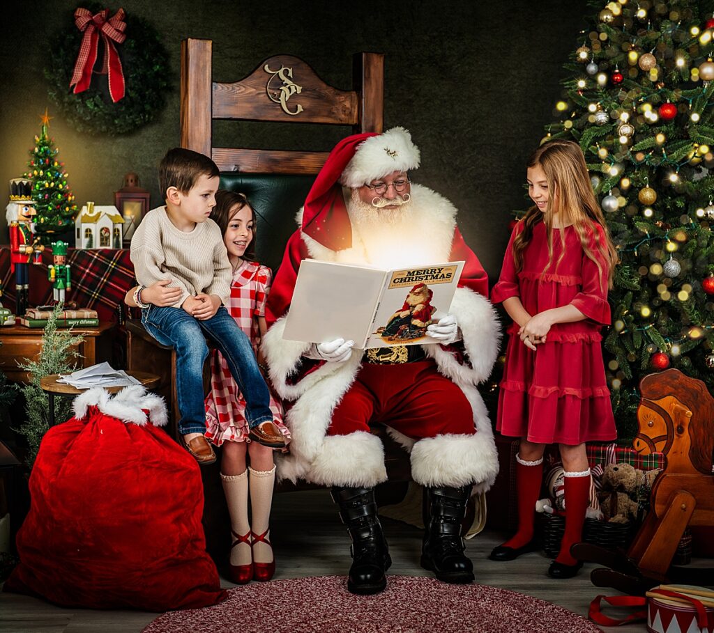 Three kids dressed in their christmas best stand around santa as he reads them a story during their santa mini session in Asheville. 