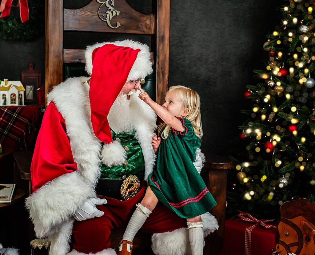 A little girl dressed in her christmas dress sits on Santa's lap and boops his nose. 