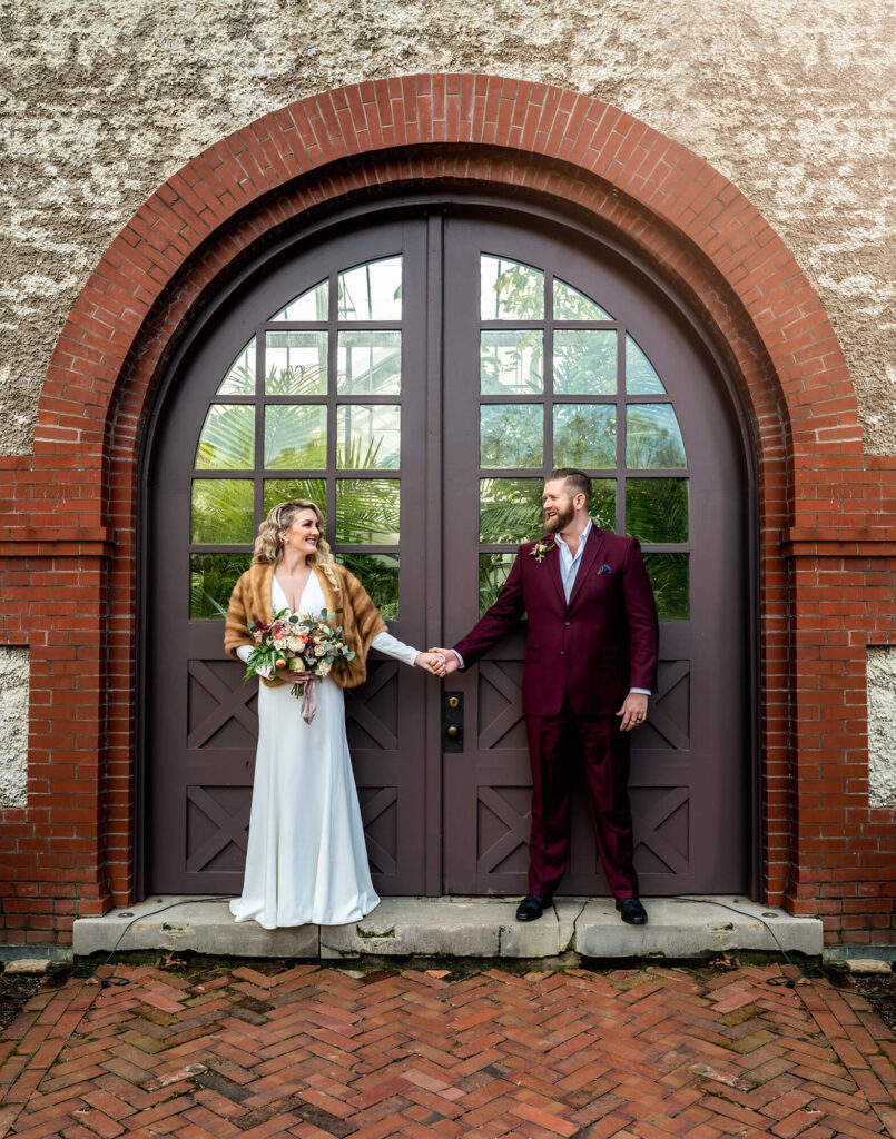 A bride and groom stand in an arched doorway and hold hands as they looking into each other's eyes. 