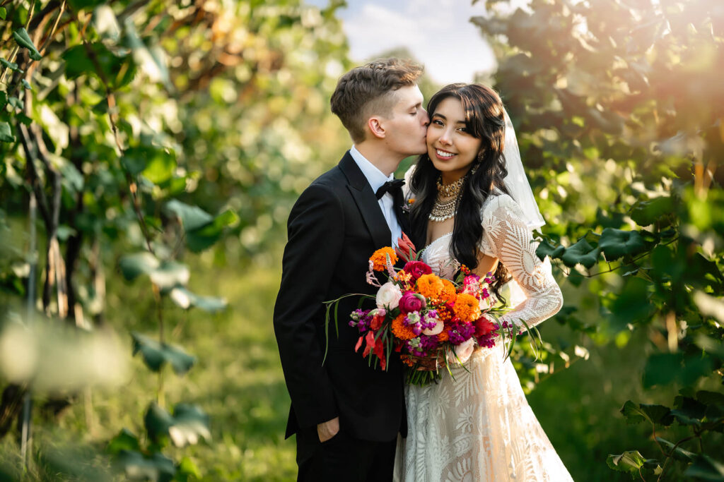 a bride and groom cuddle surrounded by vineyard vines at sunset. 