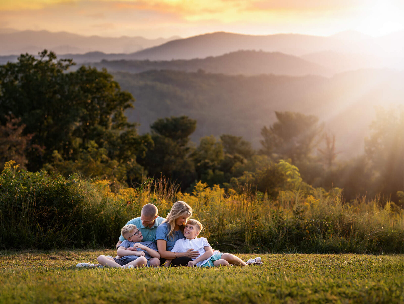 Family sitting together in a golden field with the Blue Ridge Mountains glowing at sunset near Asheville, NC