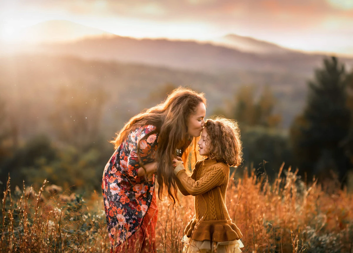 Mother and young daughter sharing a tender moment in a golden-hour field with Asheville mountains behind them