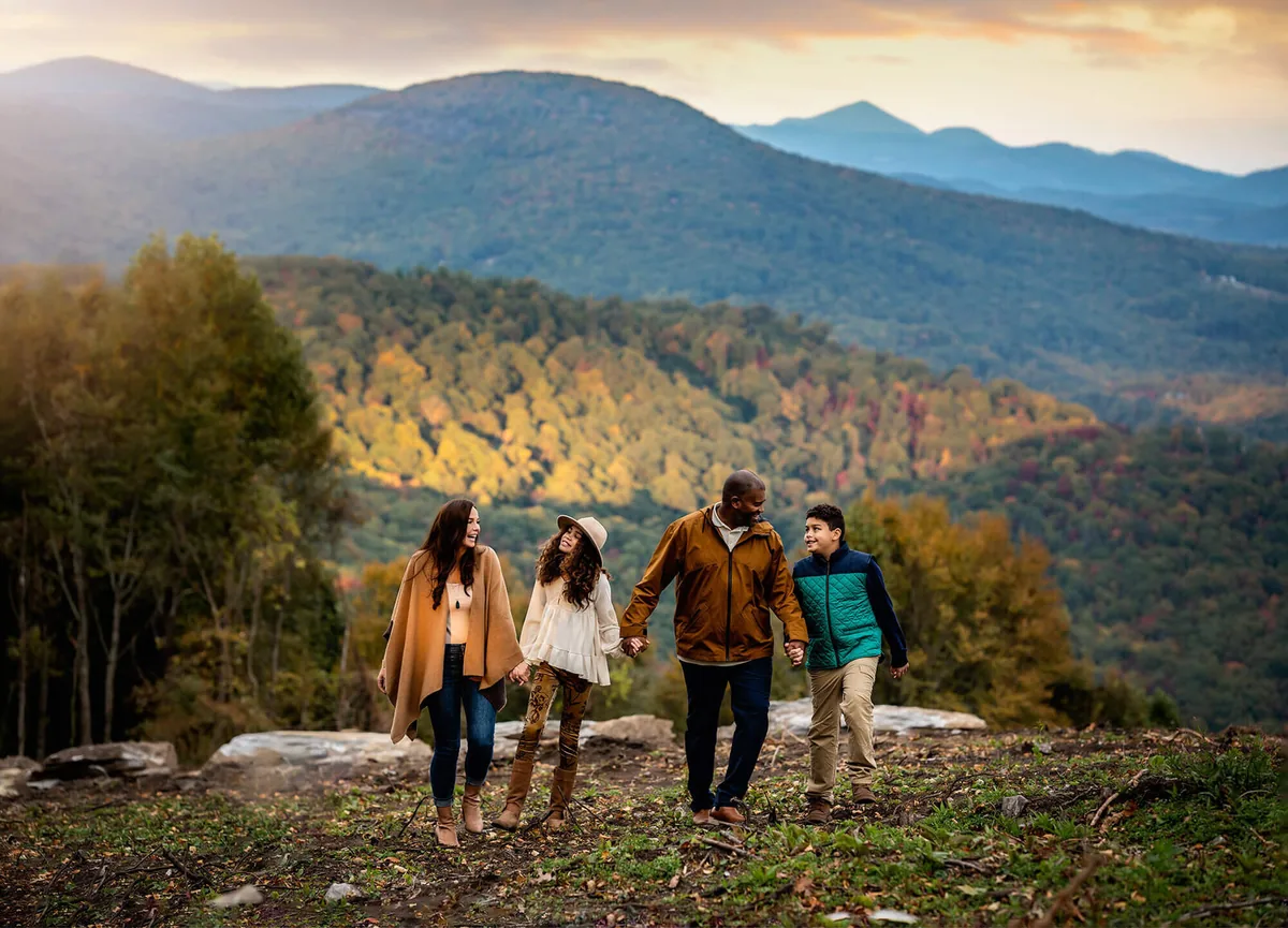 Family of four walking together on a mountain overlook with layered Blue Ridge ridgelines behind them near Asheville, NC